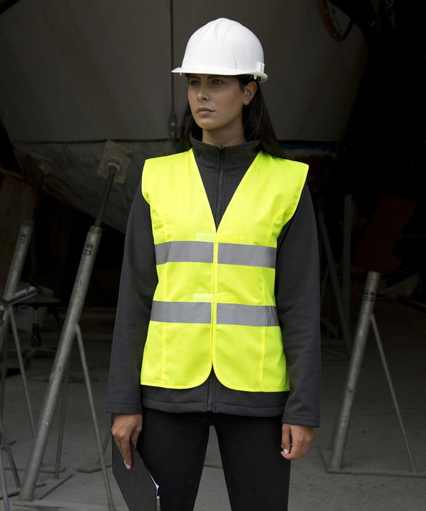 Person wearing a white hard hat and a Result R334F neon yellow safety vest stands in a construction area holding a clipboard.