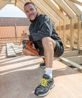 A man in a dark jacket and shorts kneels on a wooden structure holding an orange Result R348X, with an under-construction roof framework in the background.