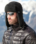 A person wearing sunglasses and a black Result R358X winter hat with ear flaps and a black puffer jacket, stands against a snowy mountain background.