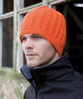 A man wearing a Result R370X orange knitted hat and black jacket stands outside, looking to the side. A weathered window is partially visible in the background.