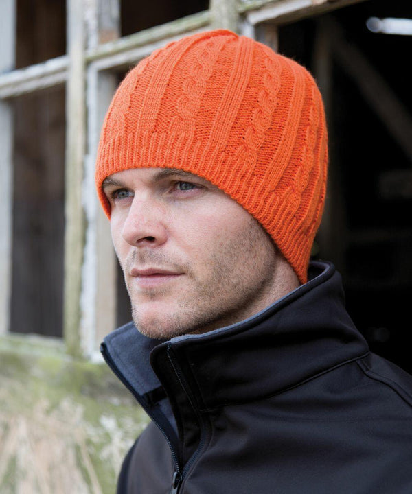 A man wearing a Result R370X orange knitted hat and black jacket stands outside, looking to the side. A weathered window is partially visible in the background.