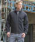 A man with blonde hair wears a Result R455M jacket and gray pants, standing in front of a construction site with scaffolding.