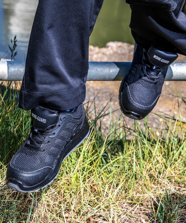Person wearing black Result R456X sneakers and black pants sits on a metal rail with grassy terrain and water in the background.
