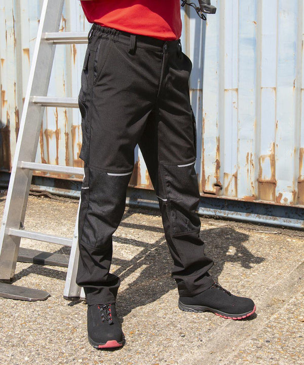 Person wearing black Result R473X work pants and black shoes stands near a metal ladder against a rusted, corrugated metal wall.