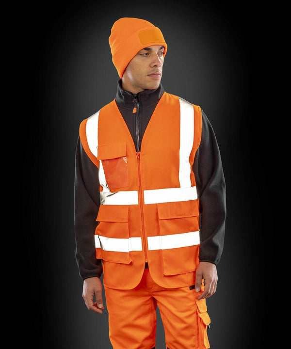 Person wearing an orange Result R477X high-visibility safety vest, orange beanie, and orange work pants, with a black long-sleeve shirt underneath, standing against a dark background.
