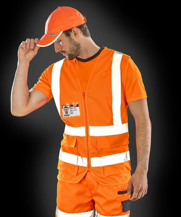 A man in an orange cap, vest, and work attire with reflective stripes adjusts his hat. He wears an ID badge on his Result R479X vest. The background is black.
