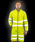 A person stands wearing the Result R505X high-visibility yellow jacket and shorts with reflective stripes against a black background.