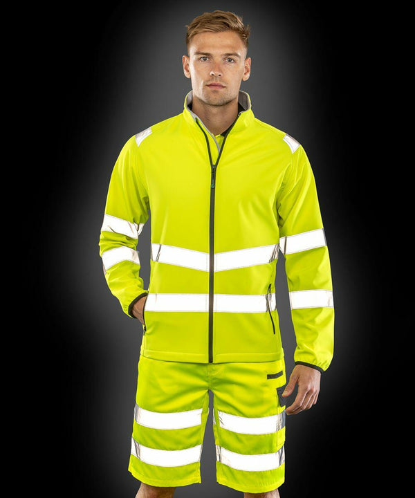 A person stands wearing the Result R505X high-visibility yellow jacket and shorts with reflective stripes against a black background.
