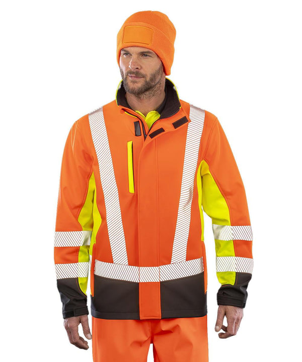 A person models the Result Recycled 3-layer printable 3-tone safety softshell, featuring an orange high-visibility jacket with reflective strips, yellow panels, black accents, a matching beanie, and orange trousers.