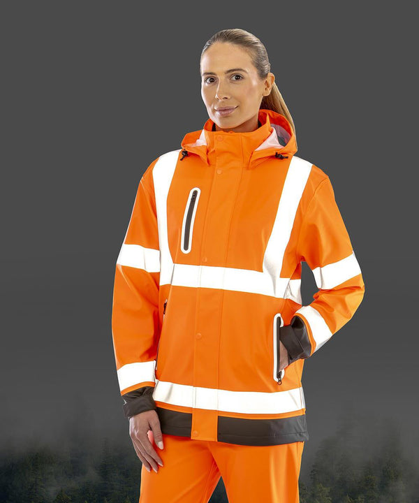 A person stands against a plain grey background wearing the Result Recycled Heavy-duty prism PU safe and dry jacket with recycled backing and matching trousers with reflective white stripes, hand in pocket, and a slight smile.