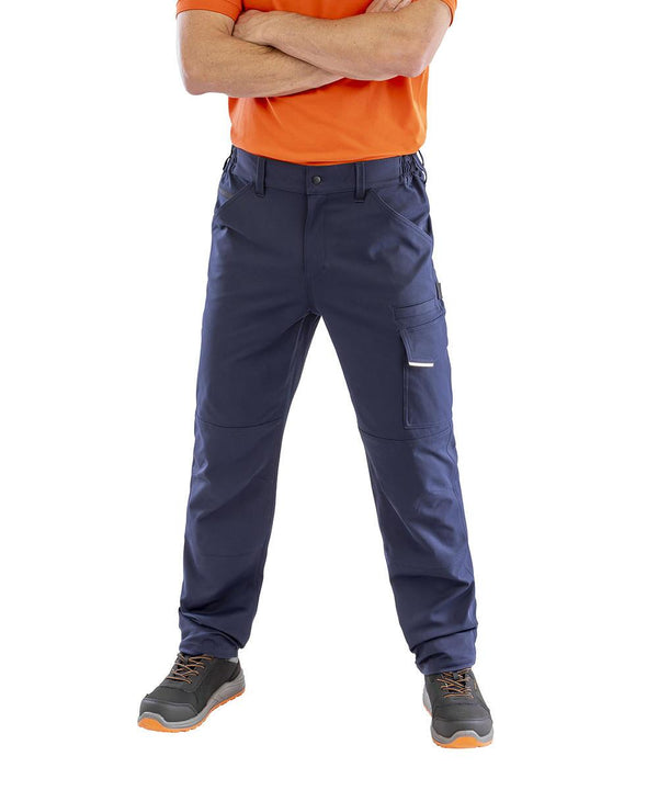 A person wearing a bright orange short-sleeve shirt, Result Workguard Work-Guard recycled dynamic stretch trousers in navy blue, and grey work shoes with orange accents stands with arms crossed, visible from shoulders to feet.