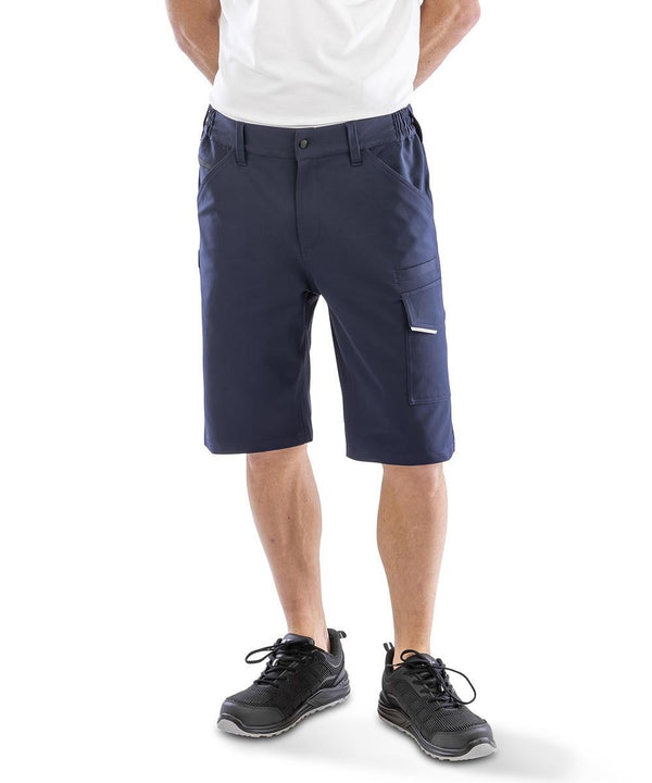 Someone stands facing forwards with hands behind their back, wearing a white T-shirt, black trainers, and Result Workguard Work-Guard recycled dynamic stretch shorts in navy blue at knee length.