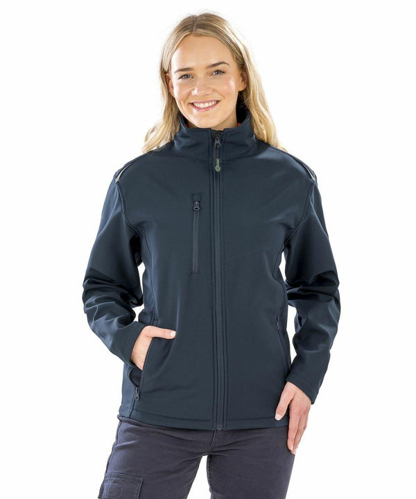 A woman wearing a dark Result Genuine Recycled R900F jacket stands with her hands in her pockets, smiling against a plain white background.
