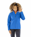 A person with curly hair wearing a blue Result R901F zip-up jacket and dark pants stands against a white background, smiling with one hand touching their hair.