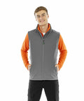 A young man wearing an orange long-sleeve shirt and a grey Result R902M smiles with his hands in his vest pockets.