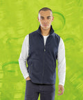 A person wearing a dark blue Result R904X vest over a white long-sleeve shirt stands against a bright green background featuring overlapping images of plastic bottles.