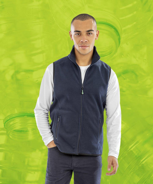 A person wearing a dark blue Result R904X vest over a white long-sleeve shirt stands against a bright green background featuring overlapping images of plastic bottles.