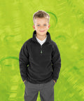 A young boy in a black Result R905J and gray pants stands with his hands in his pockets against a green background decorated with translucent bottle motifs.