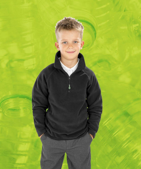 A young boy in a black Result R905J and gray pants stands with his hands in his pockets against a green background decorated with translucent bottle motifs.
