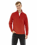 A person wearing a red long-sleeve zip-up jacket (Result Genuine Recycled R905X) and black pants stands against a plain white background, smiling with one hand in their pocket.