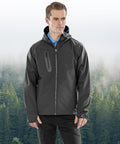 A man wearing the Result Recycled 3-layer recycled printable casual hooded softshell jacket in dark grey stands in front of a misty forest, looking slightly to the side with his hands relaxed by his sides.