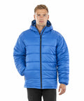 A person is wearing a Result Genuine Recycled R910X blue puffer jacket with a hood, standing against a plain white background.