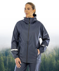 A person wears the Result Recycled Prism PU waterproof jacket with reflective bands and recycled backing, standing with one hand in a pocket against a misty forest backdrop.