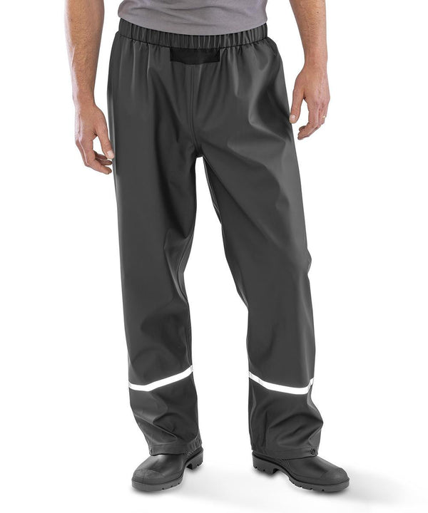 A person shown from the waist down is wearing Result Recycled Prism PU waterproof trousers with reflective bands, black boots, and a grey shirt.