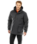 A man stands facing forward in a black Result Recycled Recycled 3-in-1 padded ripstop jacket with press-stud pockets and matte finish, paired with black trousers. The jacket is buttoned up. The background is plain white.