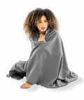 A person with curly hair is sitting on the ground, wrapped in a large gray Result Genuine Recycled R939X.