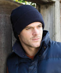 A man wearing a navy blue knit hat and a blue padded jacket from Result, model RC033, leans against a wooden wall, looking into the distance.