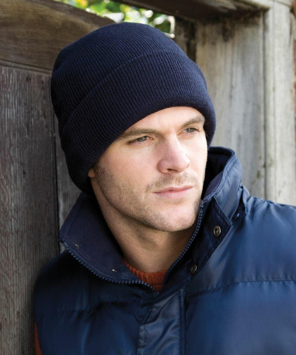 A man wearing a navy blue knit hat and a blue padded jacket from Result, model RC033, leans against a wooden wall, looking into the distance.