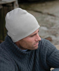 A man wearing a gray knit beanie and a thick gray sweater looks away, appearing thoughtful, while sitting outdoors near a wooden structure and a body of water. He is sporting the RC044 by Result.