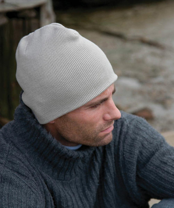 A man wearing a gray knit beanie and a thick gray sweater looks away, appearing thoughtful, while sitting outdoors near a wooden structure and a body of water. He is sporting the RC044 by Result.