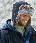A person is smiling and wearing a dark blue RC056 by Result and a fur-lined winter hat with ear flaps.