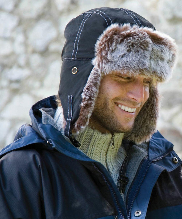 A person is smiling and wearing a dark blue RC056 by Result and a fur-lined winter hat with ear flaps.
