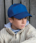 A young boy wearing a blue baseball cap, a grey hoodie, and a cream-colored knitted Result RC05J sits against a wooden fence, looking thoughtfully to the side.
