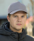 A man with short hair wears a gray Result RC05X cap and a dark jacket, looking straight at the camera with a neutral expression. The background is slightly blurred.