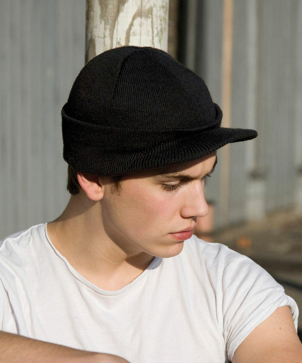 A person wearing a black knitted cap and a white t-shirt from Result, specifically the RC060 model, looks downward while sitting outdoors against a striped backdrop.