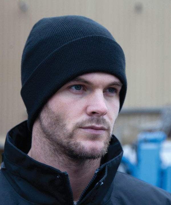 A man with a subtle beard wearing a black beanie and dark jacket looks ahead with a neutral expression. The background is blurred, showing indistinct shapes and colors.
