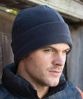 A man wearing a dark knit beanie, a black jacket, and standing in front of a wooden structure looks into the distance, sporting the Result RC141.