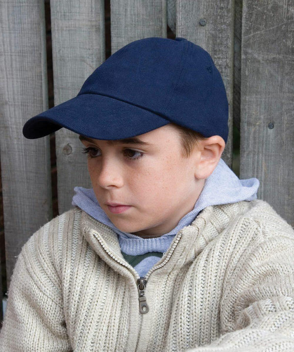A young boy wearing a navy blue cap, a gray hoodie, and a beige knit sweater sits against a wooden fence looking to the side.