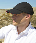 A person wearing a black RC24P Result baseball cap and white polo shirt is sitting outdoors, looking into the distance, with a wooden background visible.