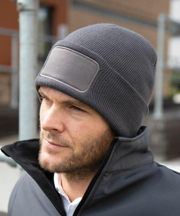 A man wearing a gray beanie and a black Result RC27X jacket stands outdoors, looking off to the side.