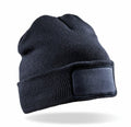 The RC27X by Result Winter Essentials is a black knit beanie featuring a blank rectangular patch on the front, shown isolated on a white background. The beanie seems to be floating, with a subtle shadow cast below it.