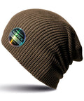 The RC31X by Result Winter Essentials is a ribbed brown beanie featuring a round, colorful patch on the side. The patch includes text and a small logo, providing a stylish enhancement to the simple knit design. It is presented against a white background in the photograph.