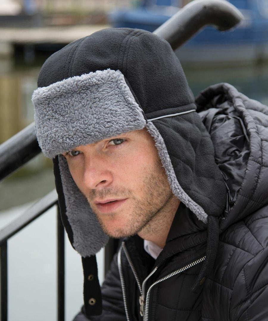 Person wearing a black winter hat with gray fleece lining and a black RC32X quilted jacket by Result, sitting near a metal railing.