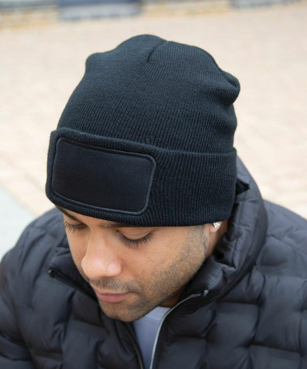 A person wearing a black beanie and a black quilted jacket outdoors. The beanie has an RC34X rectangular patch on the front from Result.