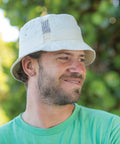 A man with a beard smiles while wearing a light green shirt and a beige Result RC45X bucket hat. Green foliage is blurred in the background.