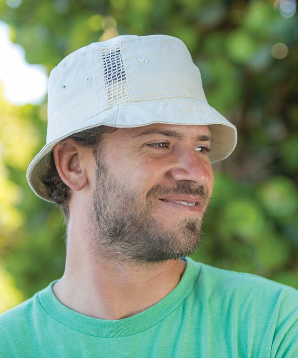 A man with a beard smiles while wearing a light green shirt and a beige Result RC45X bucket hat. Green foliage is blurred in the background.